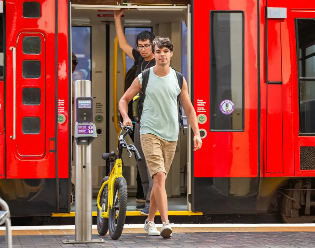 Two people with bicycles at a train station platform with red trains in the background.