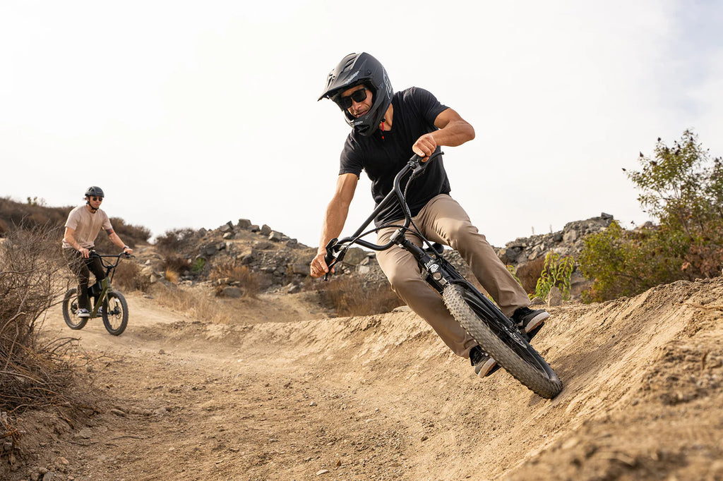 Two people riding BMX bikes on a dirt trail with a scenic background.