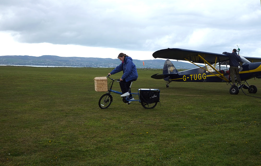 Person with a bicycle carrying a box near a small airplane on a grassy field.