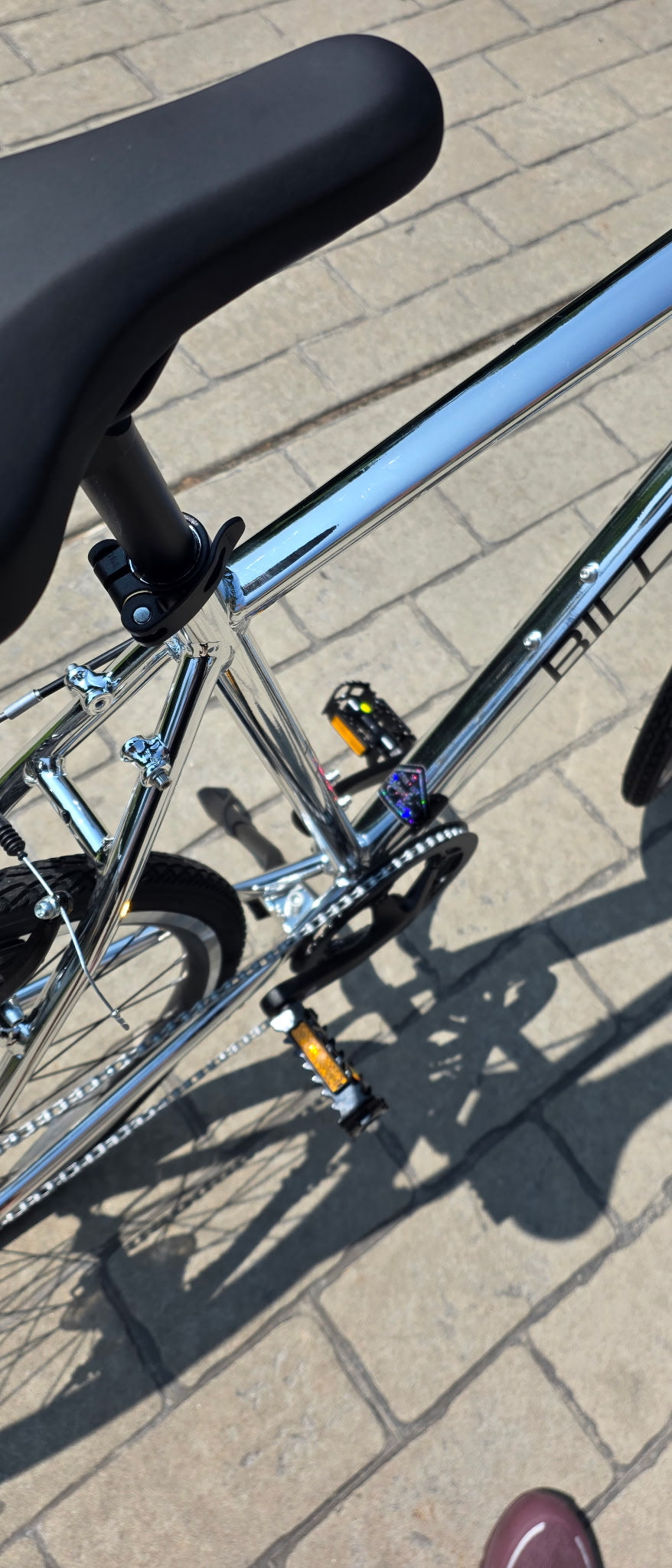 A close-up outdoor shot of a bicycle frame and saddle on a paved stone surface. The frame is metallic silver with black components, including the saddle, tires, and chainring, and small yellow accents near the pedal area.