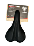 A black wide saddle bike seat with perforated details, shown with its original retail packaging on a gray wood plank background. The packaging features the WTB brand logo and 'SPEED SHE' text.